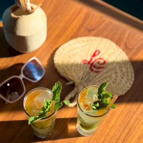 Two cocktails with mint leaves on a wooden table, a slice of melon or round bread with red text, sunglasses, and a coconut candle in the background.