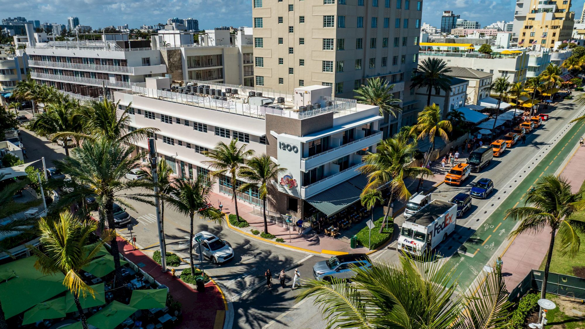 A sunny urban street with palm trees, mid‑rise buildings, parked cars, and a few buses along a seaside avenue, bright NYC-style coastal vibe.