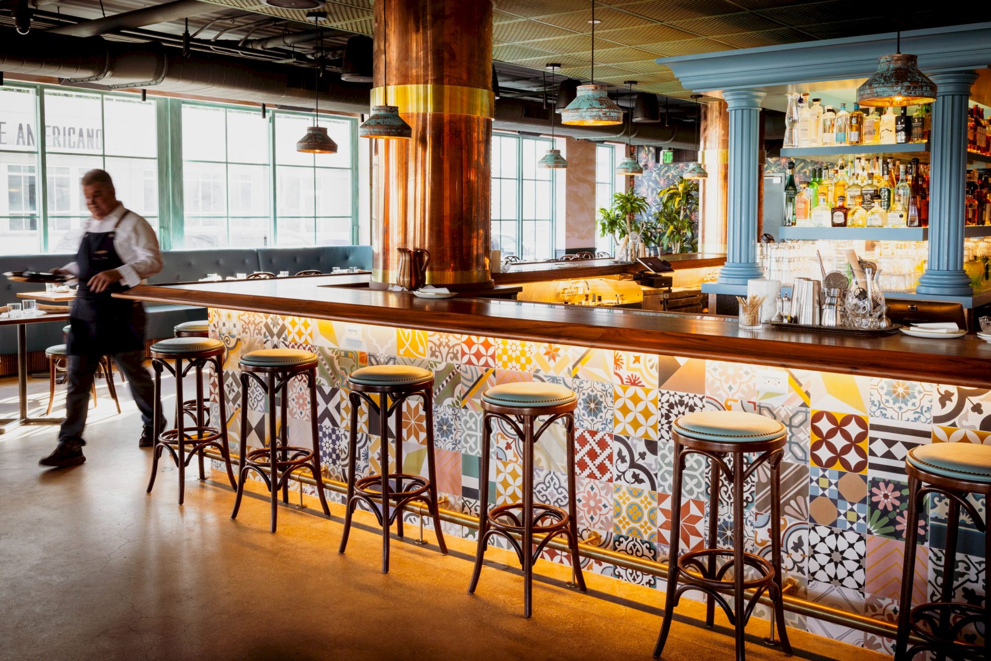 A bright, cozy bar with patterned tile counter, high stools, warm lighting, and a chef or staff member near the back.