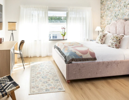 A bright, airy bedroom with a plush pink upholstered bed, floral wallpaper accent wall, sheer white curtains, light wood flooring, a desk and chair, and a patterned rug by the foot of the bed.