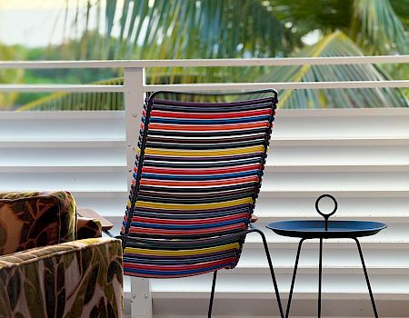 A colorful striped chair on a balcony next to a small black metal table and a cozy sofa, with tropical greenery in the background.