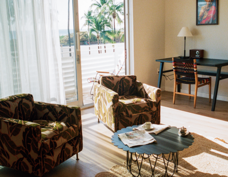 A cozy living room with two patterned armchairs, a round coffee table, a rug, and a desk by a window with sheer curtains and a sunny balcony view.