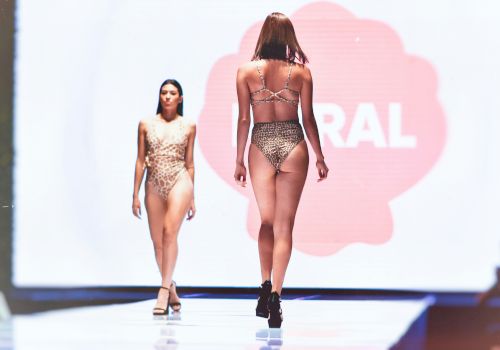 A fashion show with two models walking the runway in swimsuits, bright stage lights, and a large red circular backdrop.