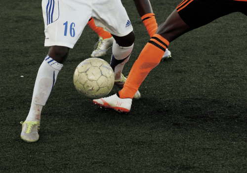 Two soccer players challenge for the ball; white and orange kits, close contact on a dark turf field, soccer cleats and ball at foot.