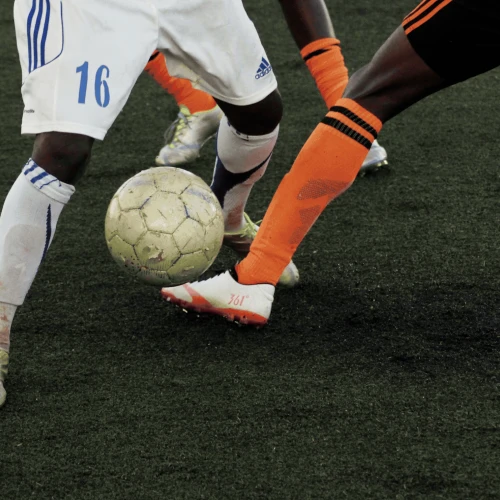 Two soccer players challenge for the ball; white and orange kits, close contact on a dark turf field, soccer cleats and ball at foot.