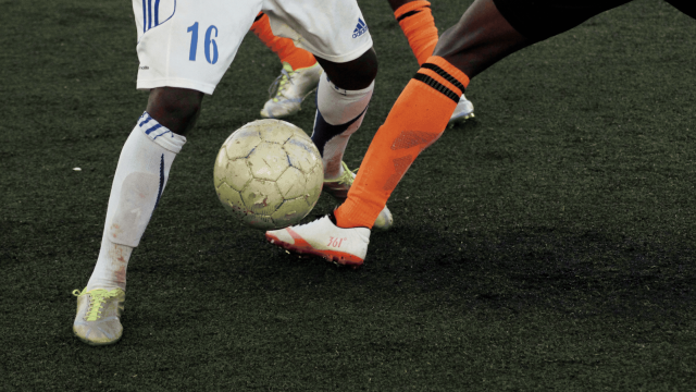 Two soccer players challenge for the ball; white and orange kits, close contact on a dark turf field, soccer cleats and ball at foot.