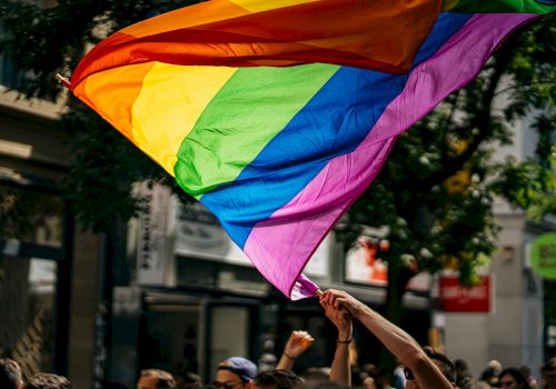 A large rainbow flag waving above a crowd at what looks like a street parade or rally, with people raising their hands in support.