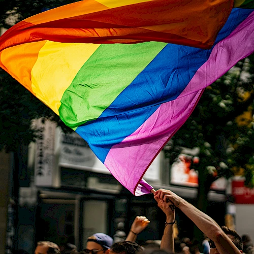 A large rainbow flag waving above a crowd at what looks like a street parade or rally, with people raising their hands in support.