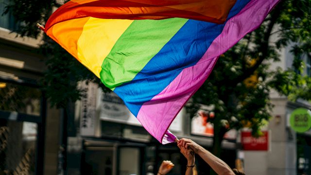 A large rainbow flag waving above a crowd at what looks like a street parade or rally, with people raising their hands in support.