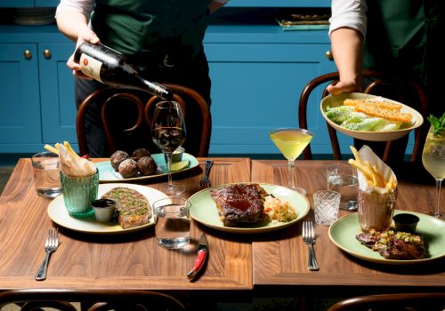Two waiters serve a fine, multi-course meal: steaks, sauces, fries, sauces, wine, and cocktails on a wooden table with blue cabinets behind.