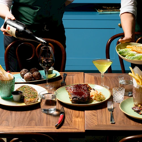Two waiters serve a fine, multi-course meal: steaks, sauces, fries, sauces, wine, and cocktails on a wooden table with blue cabinets behind.