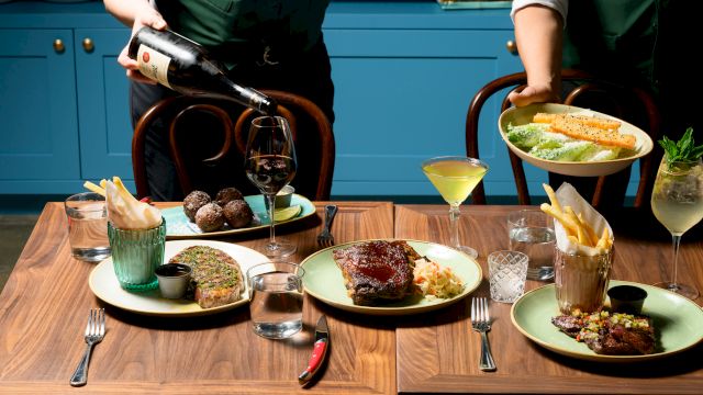 Two waiters serve a fine, multi-course meal: steaks, sauces, fries, sauces, wine, and cocktails on a wooden table with blue cabinets behind.