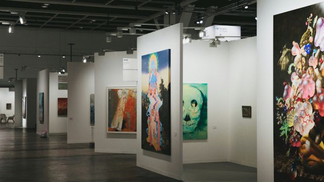 A modern art gallery with tall white partitions displaying colorful paintings on both sides, subdued lighting, and a spacious interior.