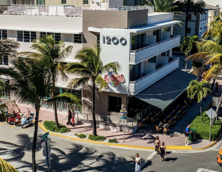 A sunny beachfront street scene with a mid-century hotel labeled &ldquo;1200&rdquo; beside palm trees, parked cars, and people walking by on the sidewalk.
