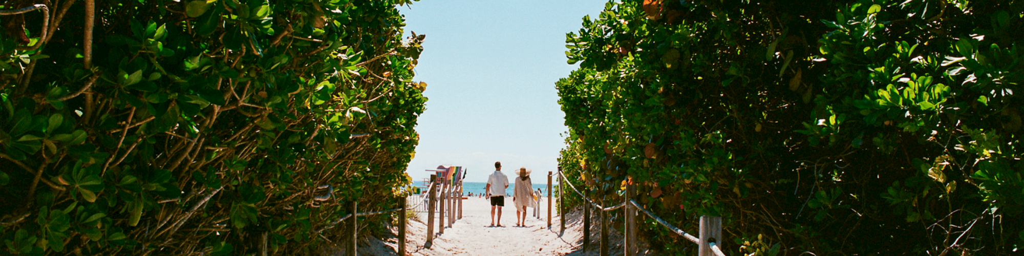 A sandy path between green shrubs leads to the beach, with a few people in the distance and a bright blue sky above.