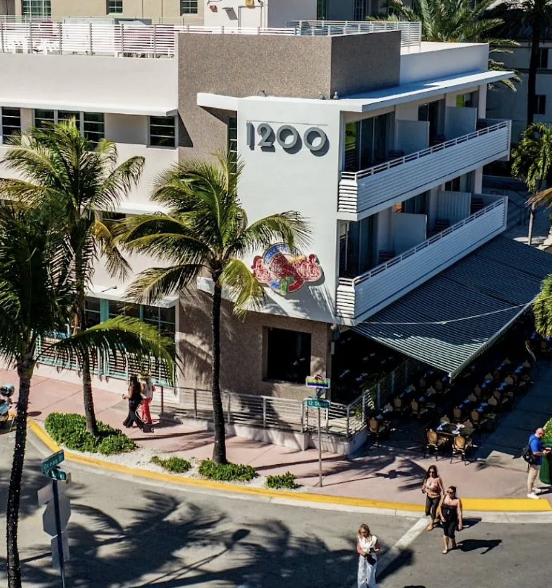 A sunny street scene at a retro hotel or cafe in a tropical town, with palm trees, people walking, and orange taxis nearby.