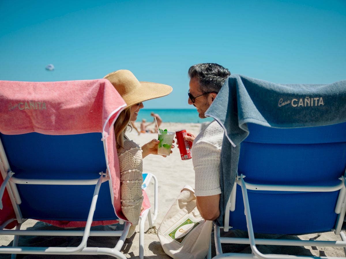 Two people sit in beach chairs facing each other, wearing hats, with drinks, towel-covered chairs, and a sunny blue sky over the sand.