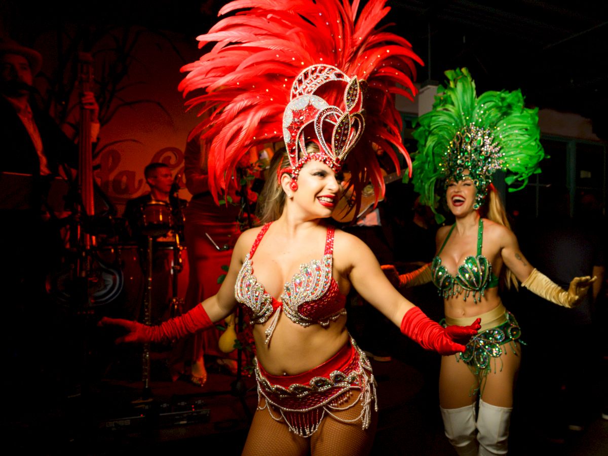 Two dancers in elaborate carnival costumes with large feather headdresses, sequins, and smiling faces performing on a dark stage.