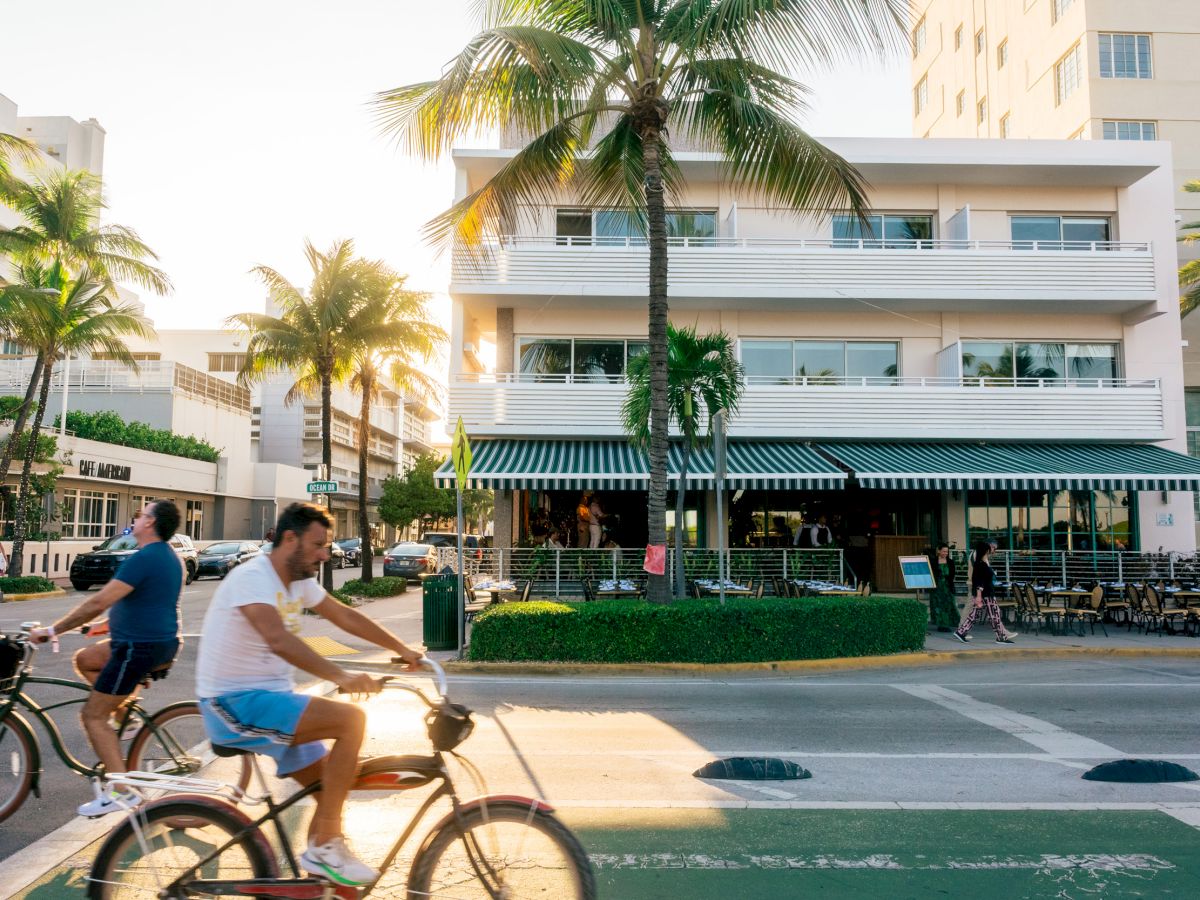 A sunny street scene with cyclists riding past a white, mid-century building, palm trees, and a cafe-front with striped awnings.