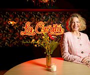 A smiling woman in a pink checkered blazer stands beside a lit round table with a small vase and candle, La Cantina sign in the background.