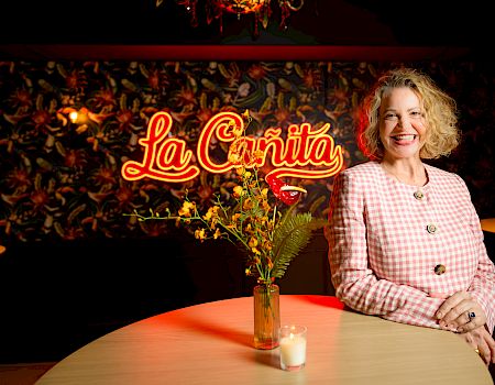 A smiling woman in a pink checkered blazer stands beside a lit round table with a small vase and candle, La Cantina sign in the background.