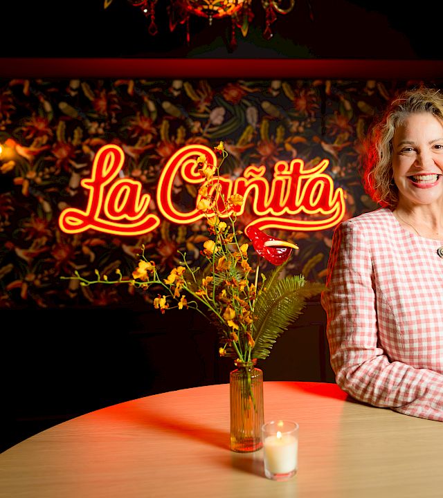 A smiling woman in a pink checkered blazer stands beside a lit round table with a small vase and candle, La Cantina sign in the background.