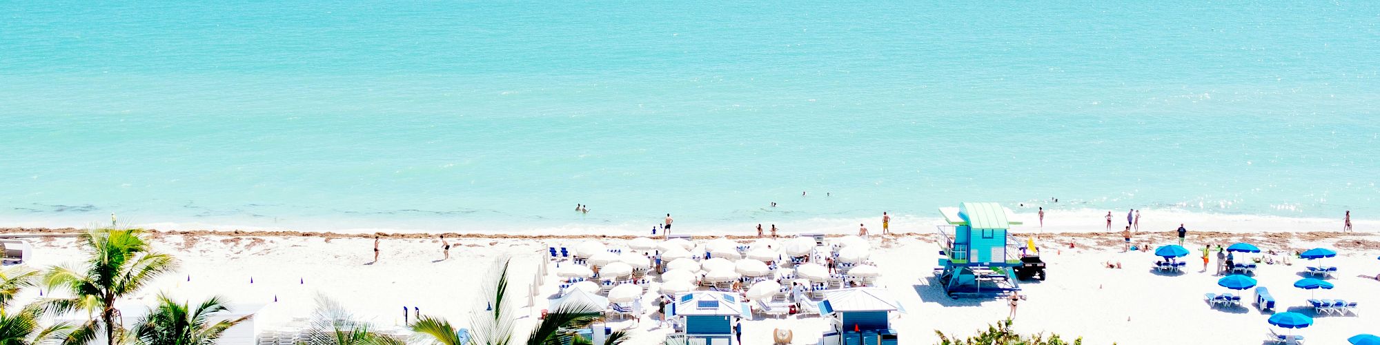 Sunny beach scene with people, umbrellas, white cabanas, and colorful chairs along a calm turquoise shoreline.