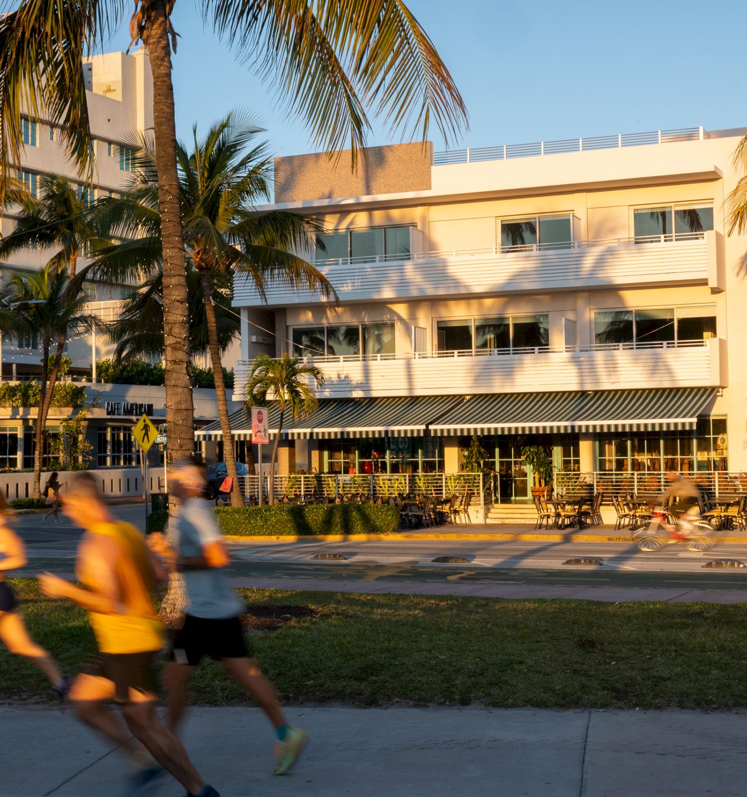 Sunlit resort courtyard with palm trees, a modern multi-story building, and people walking past outdoor seating.