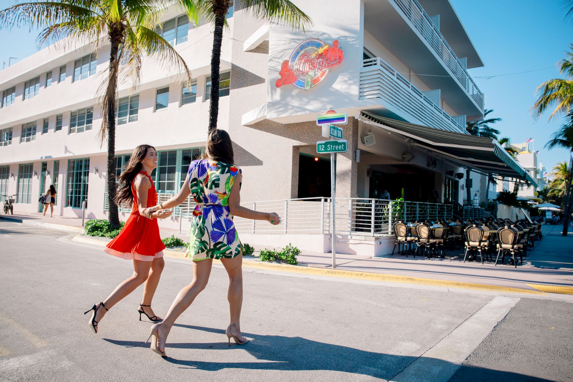 Two women stroll past palm trees and a hotel bar on a sunny street marked &ldquo;12 Street&rdquo;.