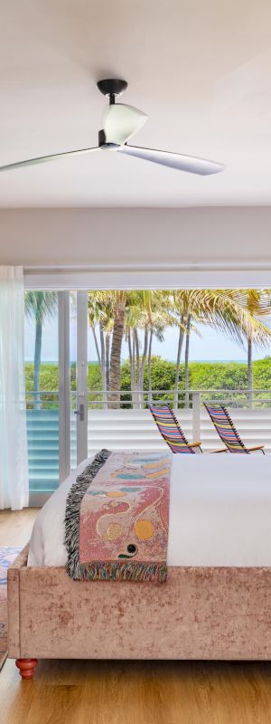 A bright bedroom with a plush bed, TV, ceiling fan, lamps, and tropical wallpaper, overlooking palm trees through large windows.