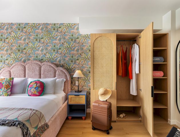 A bright modern bedroom with a plush bed, tropical patterned wallpaper, and an open wooden closet holding hanging clothes and towels.