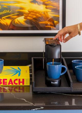 Hand placing a coffee pod into a Breville machine beside blue mugs and books, with a framed &ldquo;Year Round Paradise&rdquo; print behind.