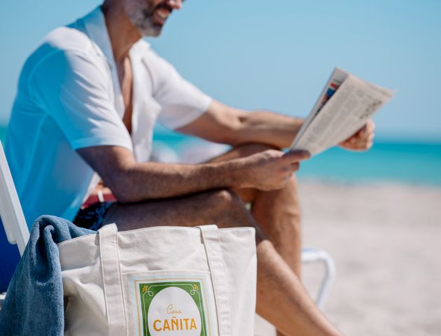 A man relaxing on a sunny beach, reading a newspaper, with tote bags and a towel nearby.
