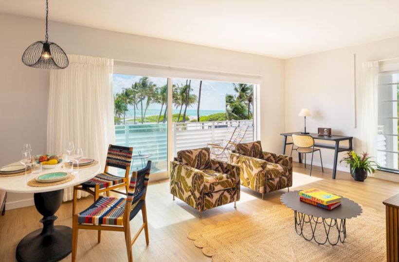 A bright coastal living room with patterned sofas and a dining set, large windows showing palm-lined ocean views.