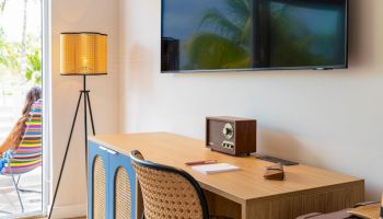 A modern home office setup with a wooden desk, rattan chairs, a lamp, a TV screen, and a small speaker on the tabletop.