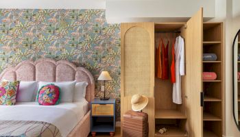 A modern bedroom with an upholstered bed, tropical patterned wallpaper, a bedside lamp, and an open wooden closet with hanging clothes.