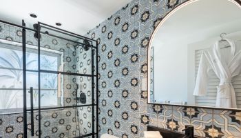 A modern bathroom with patterned mosaic tiles, a glass shower, an arched mirror, and a black countertop sink.