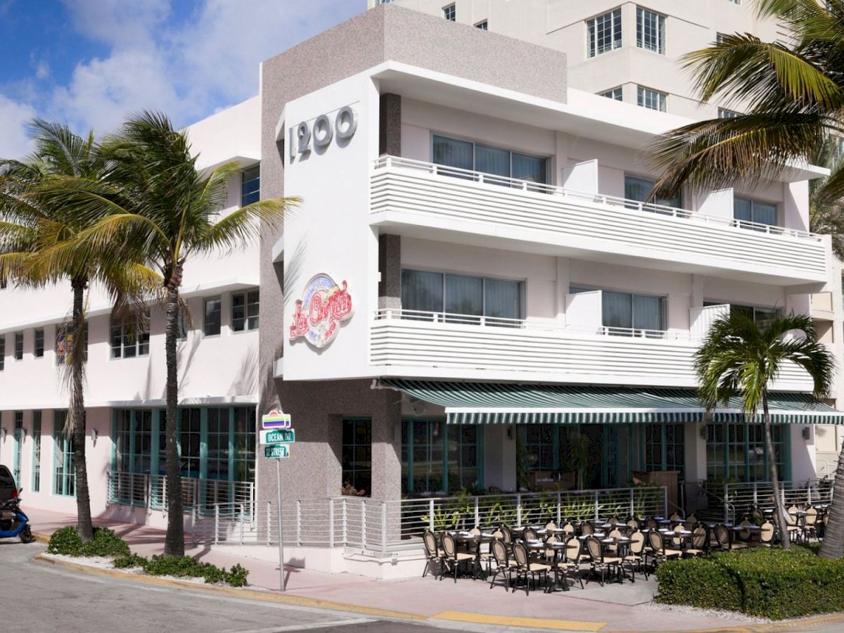 A modern hotel building with balconies and palm trees, showing signs like &ldquo;1900&rdquo; by a striped canopy.