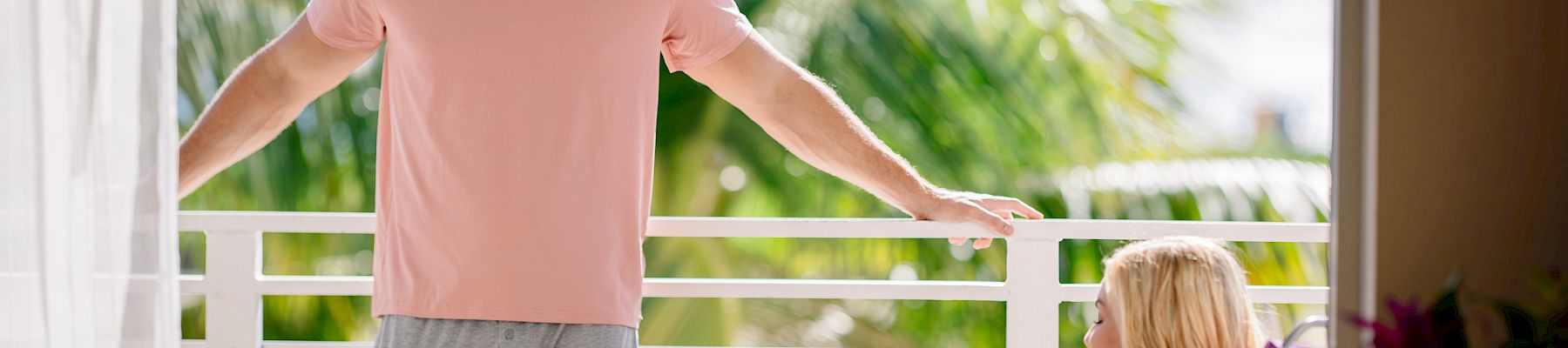 A smiling man leans on a porch rail with one arm out while a seated woman holds a cup, with tropical palm greenery blurred behind them.