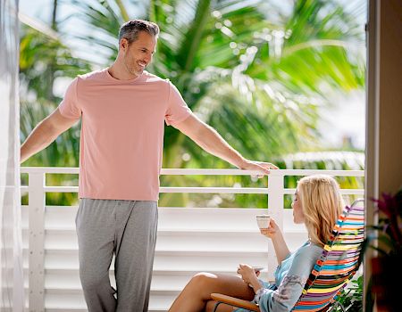 A smiling man leans on a porch rail with one arm out while a seated woman holds a cup, with tropical palm greenery blurred behind them.