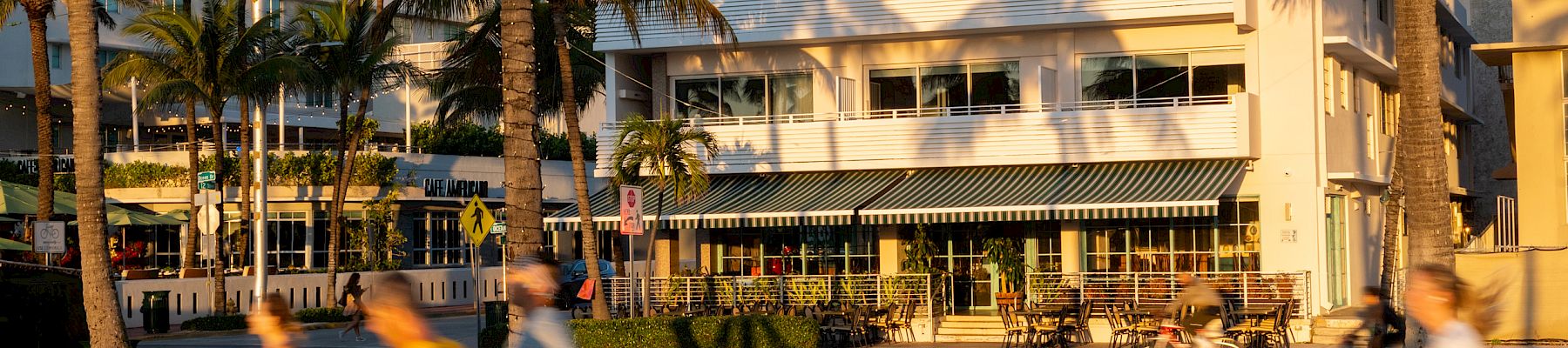 A sunny hotel courtyard with palm trees, outdoor seating, and people blurred by motion walking past.