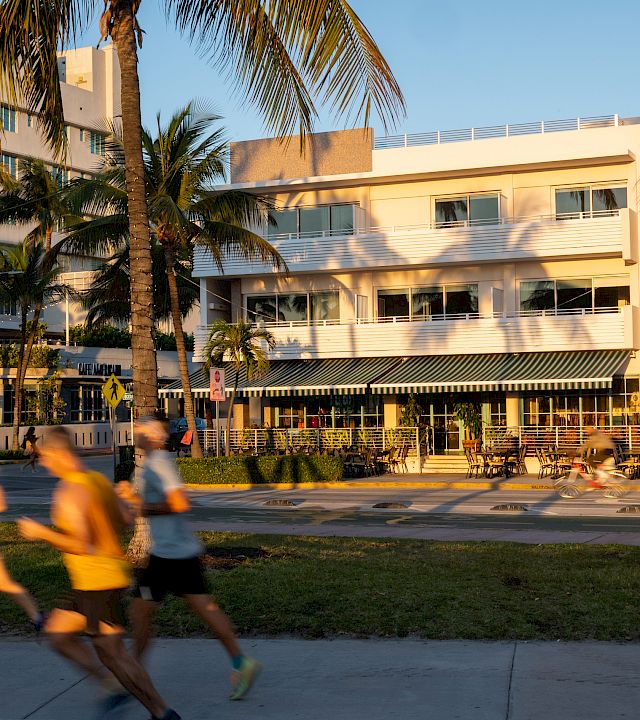 A sunny hotel courtyard with palm trees, outdoor seating, and people blurred by motion walking past.