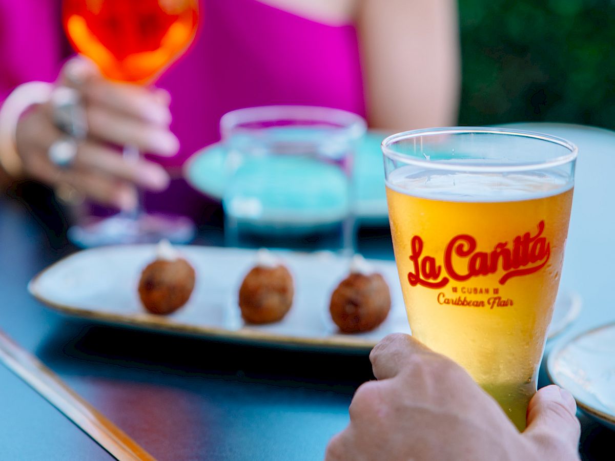 Blurry outdoor dining: a woman in a magenta top holds a small snack, with glasses and a plate of fried bites on the table.