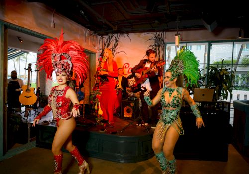 A live band plays as two costumed dancers in red and green perform on an indoor stage.