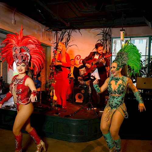 A live band plays as two costumed dancers in red and green perform on an indoor stage.
