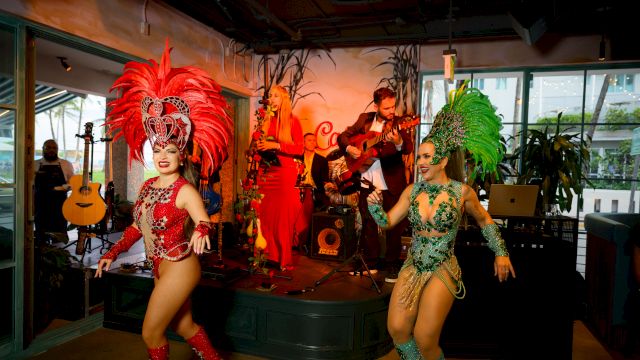 A live band plays as two costumed dancers in red and green perform on an indoor stage.