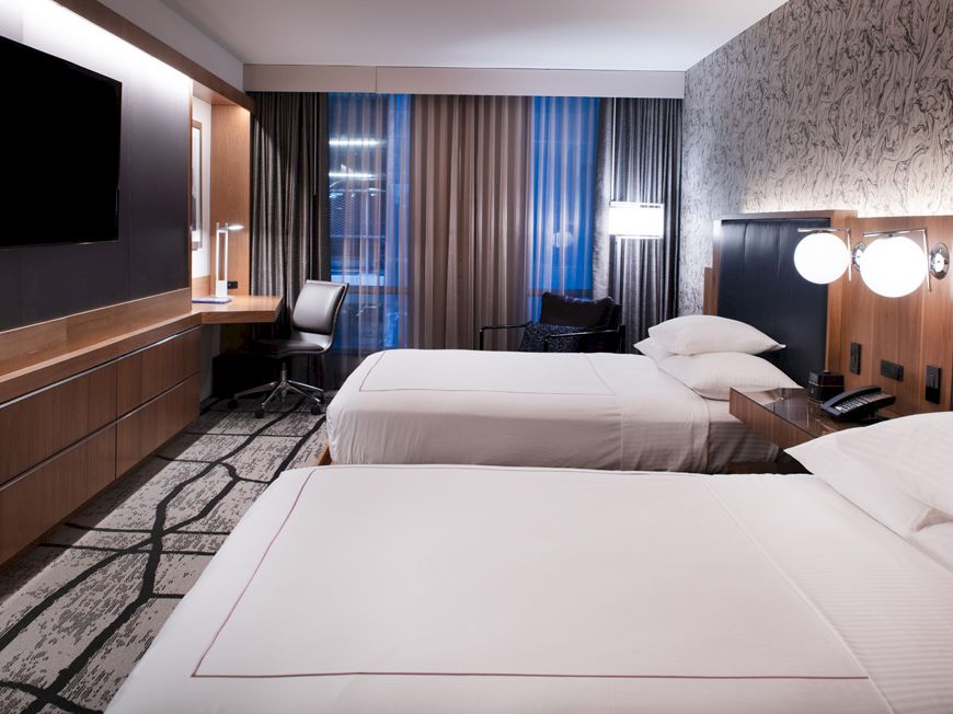 A modern hotel room with two twin beds, neutral decor, and bedside lamps with curtains behind.