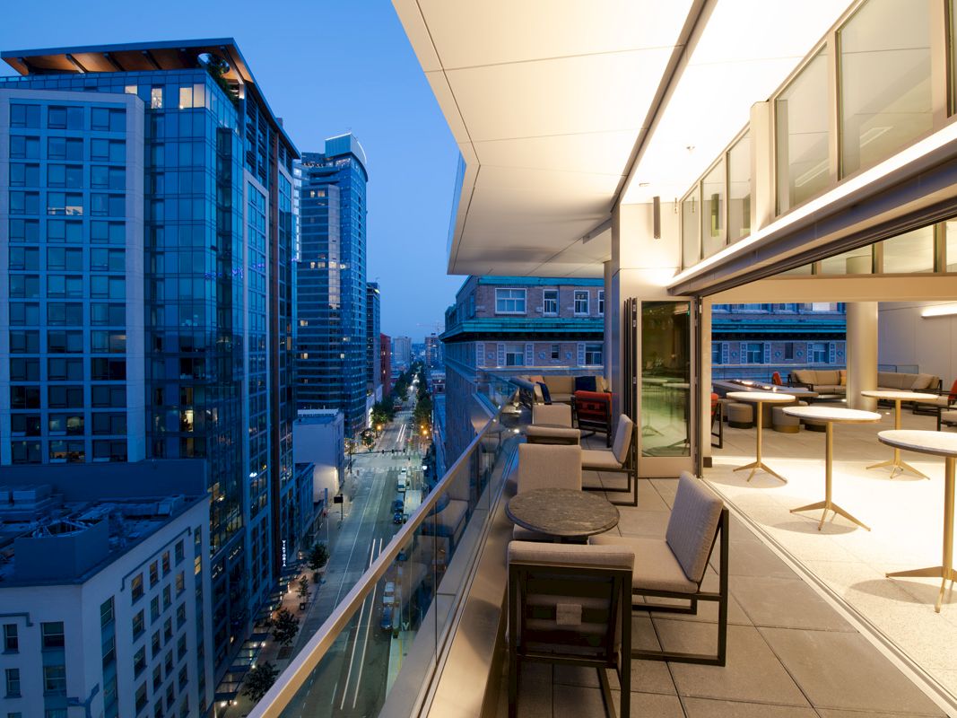 View from a modern high-rise balcony over a busy city street, with patio chairs and tables in the foreground.